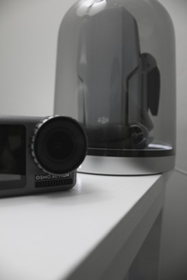 A small camera labeled 'OSMO ACTION' is placed on a white surface. Behind it, a large gray and black drone is displayed under a clear glass dome. The scene is minimalistic with a focus on technology accessories.
