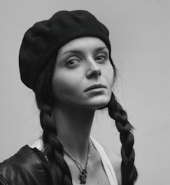 A portrait of a person wearing a dark beret and a jacket with braided hair. The expression on their face is serious and contemplative. The image is in black and white, emphasizing shadows and highlights.