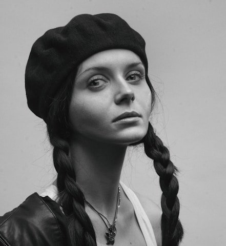 A portrait of a person wearing a dark beret and a jacket with braided hair. The expression on their face is serious and contemplative. The image is in black and white, emphasizing shadows and highlights.
