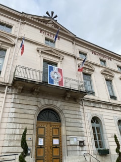 A grand historic building with 'ÉGALITÉ' and 'FRATERNITÉ' carved above the entrance, featuring a large wooden door, arched windows, and balconies. The building is adorned with three French flags and a colorful banner with a portrait and the words 'LIBERTÉ EGALITÉ FRATERNITÉ'.