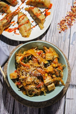 A colorful bowl of stir-fried vegetables and tofu topped with sesame seeds. Nearby, there is a plate of golden-brown dumplings drizzled with a spicy red sauce and garnished with black sesame seeds. The presentation is appetizing and vibrant, with a sprinkle of red pepper flakes scattered on the wooden table surface.