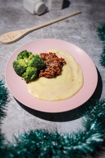 A pink plate holds a serving of creamy mashed potatoes accompanied by steamed broccoli and a portion of minced meat in a sauce. The background includes a wooden spoon marked with text and two small white containers, possibly for salt and pepper. Green tinsel decorates the lower part of the image, adding a festive touch to the gray surface.