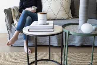 A living room setup featuring a gray sofa with a large textured cushion. In front of the sofa, there are two circular tables. The left table has a white surface displaying a candle labeled 'BROOKE' and stacked books. The other table is green with a gray textured top, holding a tall vase and round decorative objects. To the left, a person in jeans and a black top holds a cup while seated barefoot on the sofa.