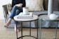 A living room setup featuring a gray sofa with a large textured cushion. In front of the sofa, there are two circular tables. The left table has a white surface displaying a candle labeled 'BROOKE' and stacked books. The other table is green with a gray textured top, holding a tall vase and round decorative objects. To the left, a person in jeans and a black top holds a cup while seated barefoot on the sofa.