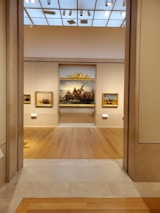 A well-lit art gallery features several framed paintings hung on beige walls. The focus is a large, prominent painting at the center, depicting a historical scene, possibly involving figures in a dramatic setting. The gallery has wooden floors and a high ceiling with spotlights.
