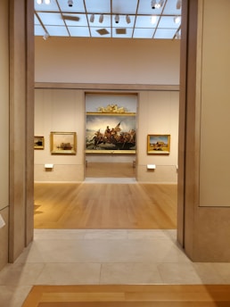 A well-lit art gallery features several framed paintings hung on beige walls. The focus is a large, prominent painting at the center, depicting a historical scene, possibly involving figures in a dramatic setting. The gallery has wooden floors and a high ceiling with spotlights.