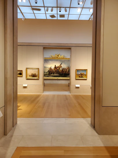 A well-lit art gallery features several framed paintings hung on beige walls. The focus is a large, prominent painting at the center, depicting a historical scene, possibly involving figures in a dramatic setting. The gallery has wooden floors and a high ceiling with spotlights.
