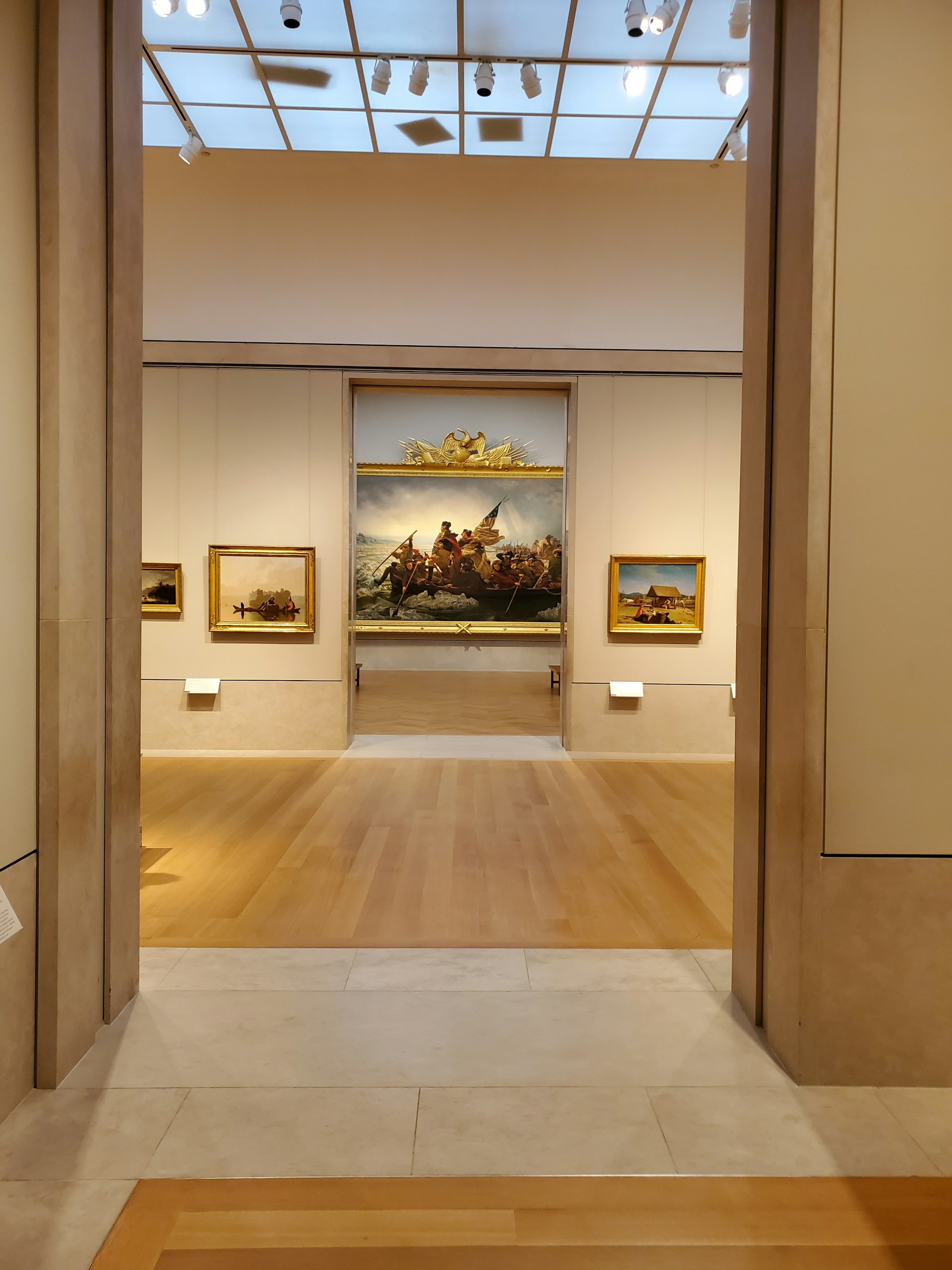 A well-lit art gallery features several framed paintings hung on beige walls. The focus is a large, prominent painting at the center, depicting a historical scene, possibly involving figures in a dramatic setting. The gallery has wooden floors and a high ceiling with spotlights.
