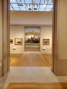 A well-lit art gallery features several framed paintings hung on beige walls. The focus is a large, prominent painting at the center, depicting a historical scene, possibly involving figures in a dramatic setting. The gallery has wooden floors and a high ceiling with spotlights.