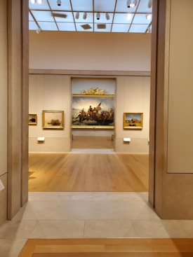 A well-lit art gallery features several framed paintings hung on beige walls. The focus is a large, prominent painting at the center, depicting a historical scene, possibly involving figures in a dramatic setting. The gallery has wooden floors and a high ceiling with spotlights.