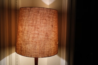 A warmly lit lamp with a textured, brown cylindrical shade is positioned against a wall, casting soft shadows. The ambient lighting creates a cozy and inviting atmosphere.