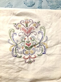 A detailed embroidery design featuring two symmetrical birds facing each other, surrounded by an intricate arrangement of floral and botanical patterns. The embroidery is colorful, with a variety of threads used to create a vibrant and elegant appearance. The background is a white fabric.