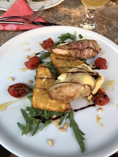 A white plate contains a gourmet dish featuring grilled chicken wrapped in bacon topped with melted cheese, accompanied by roasted cherry tomatoes and crispy potato pieces. The plate is garnished with arugula, drizzle of balsamic glaze, and scattered pine nuts. A glass of beverage is visible at the top right.