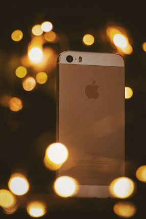 A close-up shot of a silver smartphone standing vertically against a backdrop of warm, defocused lights. The composition creates a festive and dreamy atmosphere with blurred circles of light surrounding the phone.