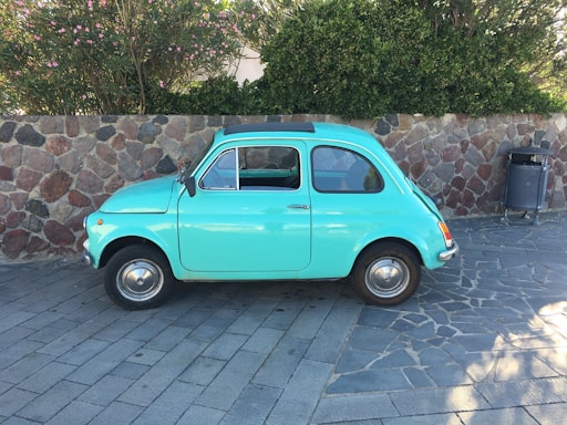 A vintage turquoise car is parked on a stone-paved road beside a stone wall. The background includes lush green foliage and flowering bushes. There is also a trash bin on the right side, near the wall.