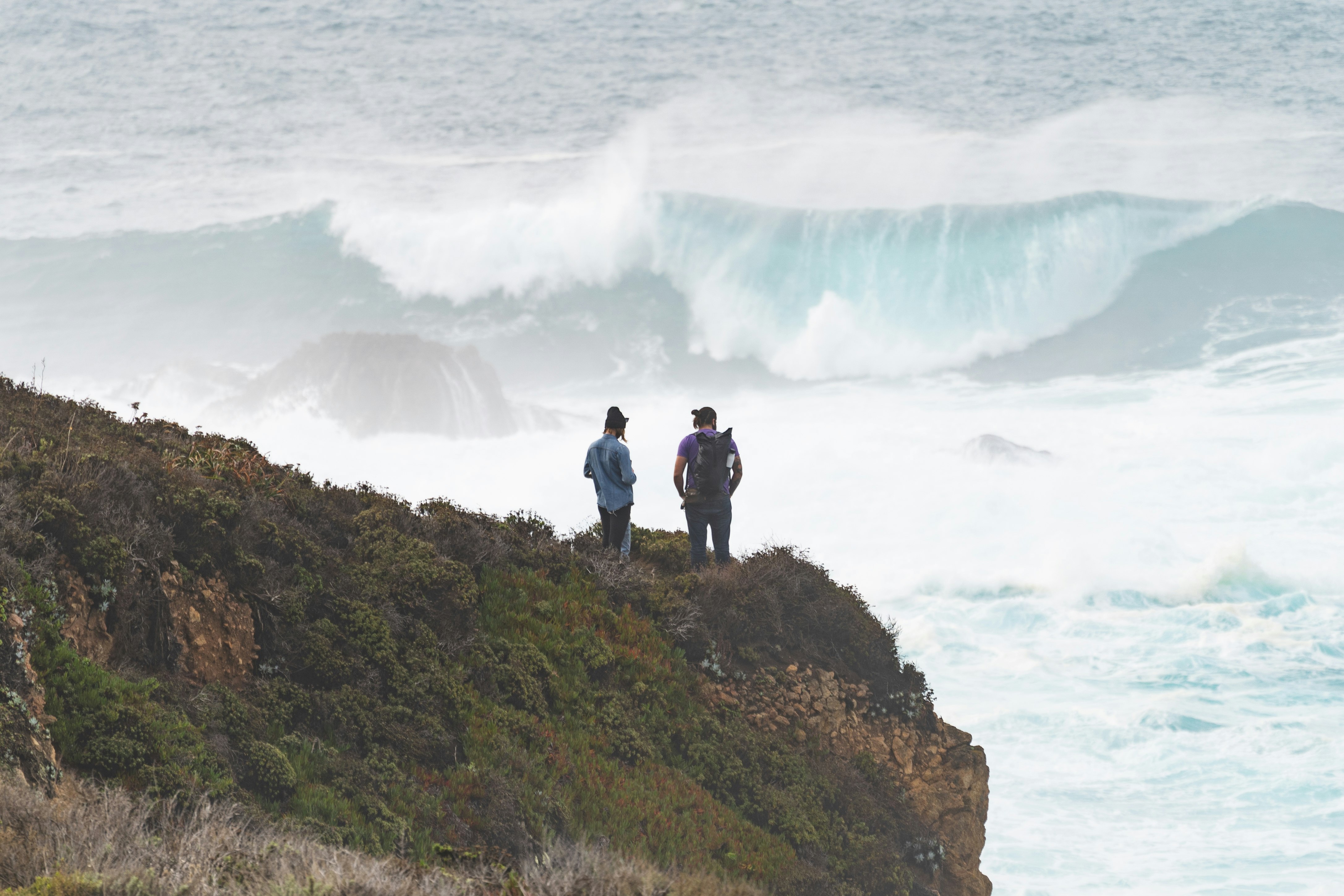 2 men standing on green grass covered hill near sea waves crashing on shore during daytime