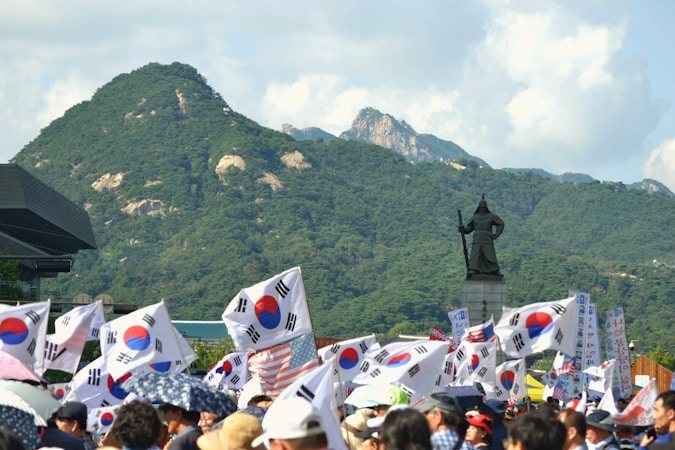 A large gathering of people is seen waving South Korean flags in front of a mountainous landscape. At the center stands a statue of a historical figure, possibly a military leader. The crowd is lively, and the scene is set under a bright sunny sky.