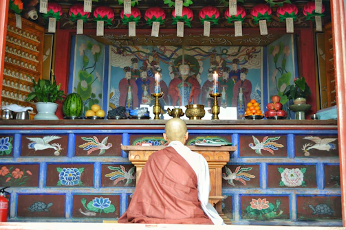 A person in traditional attire is kneeling in front of an ornate altar, which is adorned with colorful murals and several offerings including fruits and incense. The altar features intricate designs with flowers and birds, and is illuminated by lit candles.