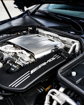 The image features the engine bay of a high-performance vehicle, showcasing a clean and well-maintained engine. Prominent branding includes an AMG logo on the engine cover, suggesting it is a Mercedes-AMG engine. The metallic components are arranged neatly, with visible pipes, wires, and textured areas.