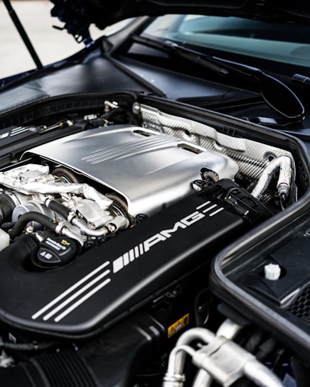 The image features the engine bay of a high-performance vehicle, showcasing a clean and well-maintained engine. Prominent branding includes an AMG logo on the engine cover, suggesting it is a Mercedes-AMG engine. The metallic components are arranged neatly, with visible pipes, wires, and textured areas.