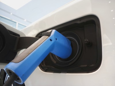 A close-up view of an electric vehicle charging port with a blue charging plug inserted into it. The surrounding area is sleek and modern, demonstrating a clean design with reflections on the white surface of the vehicle.