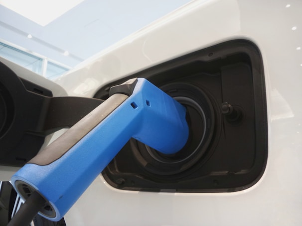 A close-up view of an electric vehicle charging port with a blue charging plug inserted into it. The surrounding area is sleek and modern, demonstrating a clean design with reflections on the white surface of the vehicle.