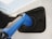 A close-up view of an electric vehicle charging port with a blue charging plug inserted into it. The surrounding area is sleek and modern, demonstrating a clean design with reflections on the white surface of the vehicle.