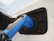 A close-up view of an electric vehicle charging port with a blue charging plug inserted into it. The surrounding area is sleek and modern, demonstrating a clean design with reflections on the white surface of the vehicle.