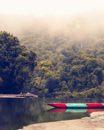 A colorful boat rests on a calm river, reflecting the misty forested hills in the background. The scene is enveloped in a gentle mist, creating a serene and tranquil atmosphere. The lush greenery contrasts with the vibrant colors of the boat, adding depth to the scenic view.
