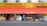 The storefront of a supermarket with a bright yellow and red awning has a variety of fruit and vegetable boxes outside. Several people are walking past the store, and the signage indicates the sale of various international products.