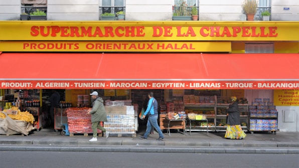 The storefront of a supermarket with a bright yellow and red awning has a variety of fruit and vegetable boxes outside. Several people are walking past the store, and the signage indicates the sale of various international products.