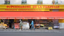 The storefront of a supermarket with a bright yellow and red awning has a variety of fruit and vegetable boxes outside. Several people are walking past the store, and the signage indicates the sale of various international products.