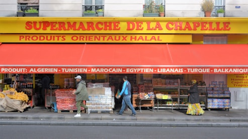 The storefront of a supermarket with a bright yellow and red awning has a variety of fruit and vegetable boxes outside. Several people are walking past the store, and the signage indicates the sale of various international products.