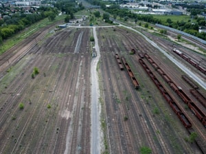 An aerial view of an expansive railway yard featuring numerous parallel train tracks. Rusty freight cars are lined up on some tracks, while others appear empty. Sparse vegetation grows between the tracks, and small buildings are visible in the background. In the distance, there are industrial buildings and residential areas, with green trees surrounding the railway.