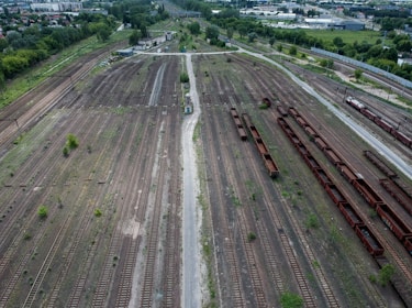 An aerial view of an expansive railway yard featuring numerous parallel train tracks. Rusty freight cars are lined up on some tracks, while others appear empty. Sparse vegetation grows between the tracks, and small buildings are visible in the background. In the distance, there are industrial buildings and residential areas, with green trees surrounding the railway.