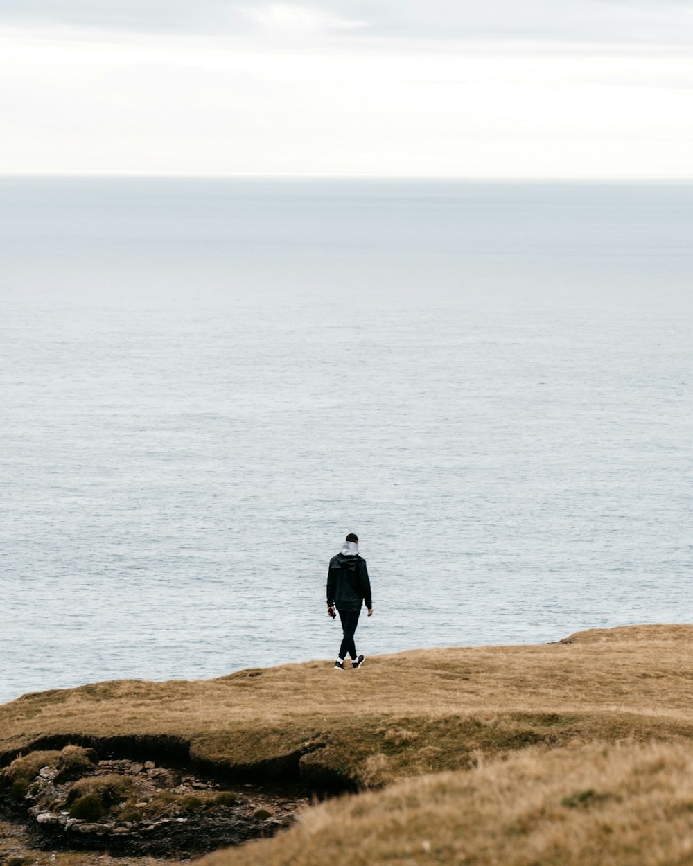 昼間、水辺の茶色の岩の上に立つ黒いジャケットを着た男