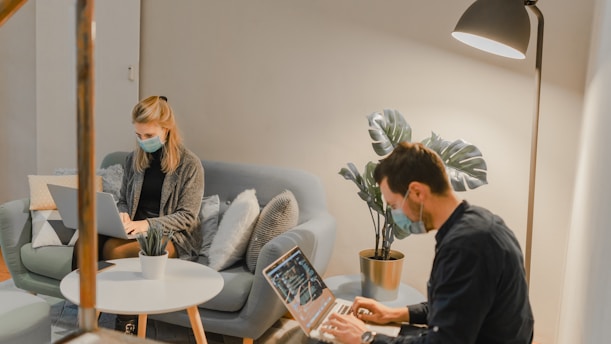 A modern indoor setting with two individuals sitting on a couch and chair, both wearing masks and working on laptops. A small round coffee table with a potted plant is placed between them. The room is softly lit with a floor lamp, and there are decorative cushions on the couch.