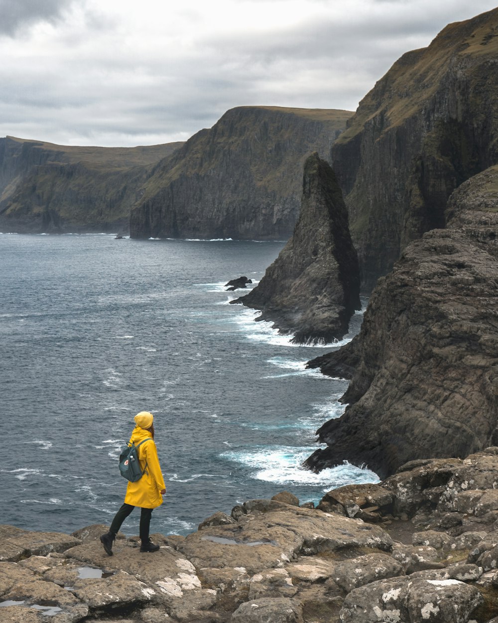 黄色いジャケットと黒いズボンを着た男が、水域近くの茶色の岩層の上に立っている