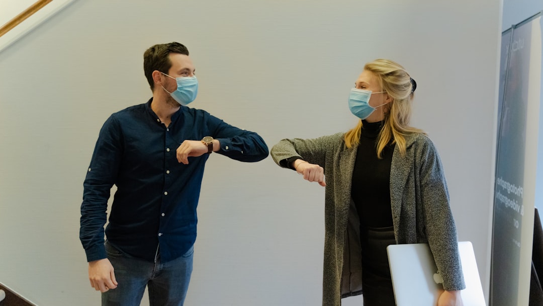Two business associates wearing masks touching elbow to elbow while smiling at each other.