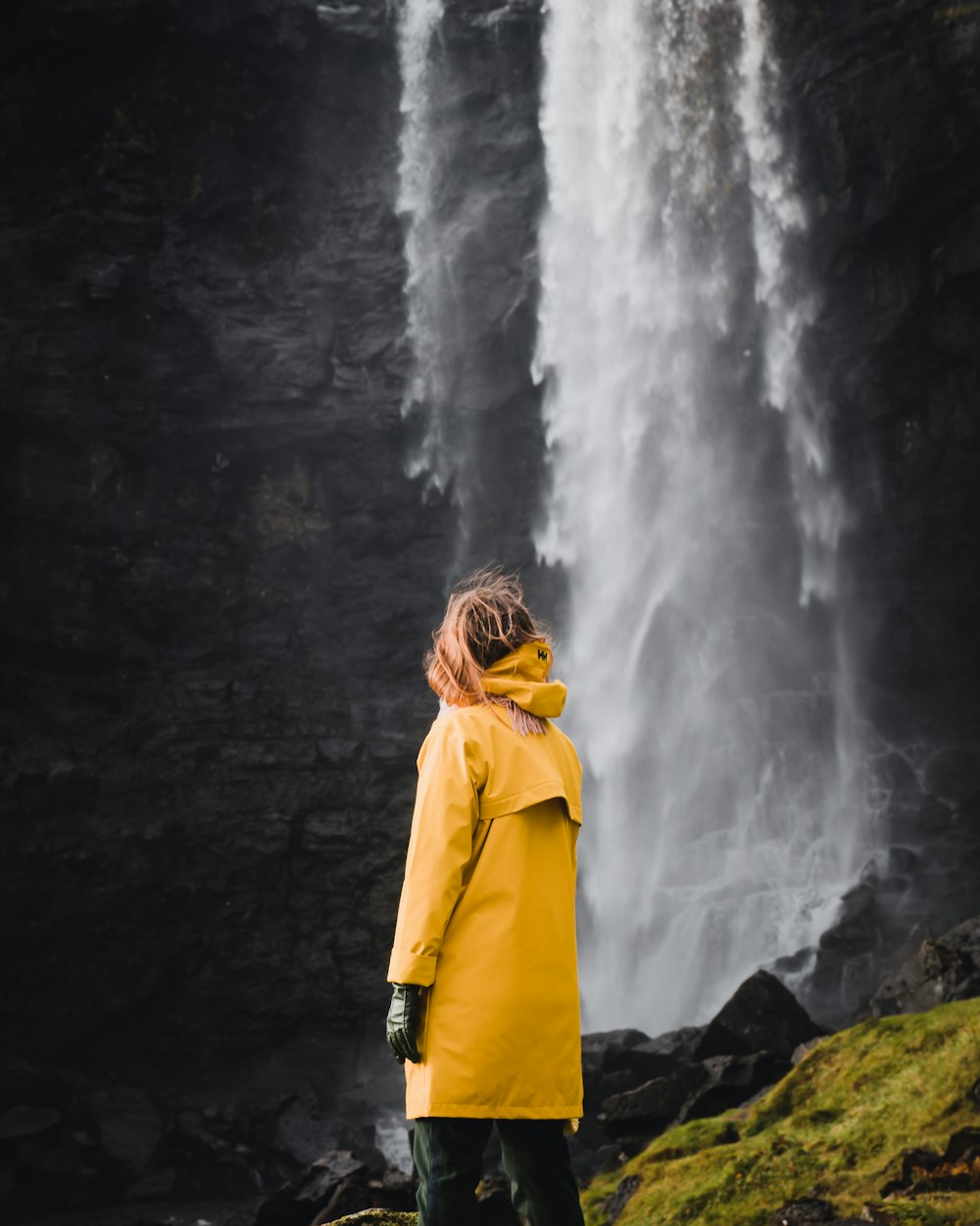 woman in yellow hoodie standing on rock near waterfalls during daytime