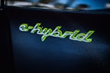 The image shows the word 'e-hybrid' written in a stylized script with green accents on a dark background. The text appears glossy and reflective, suggesting it is part of a logo or badge, possibly on a vehicle.