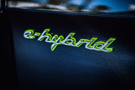 The image shows the word 'e-hybrid' written in a stylized script with green accents on a dark background. The text appears glossy and reflective, suggesting it is part of a logo or badge, possibly on a vehicle.