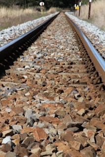 Railway tracks extend into the distance, flanked by coarse gravel stones. The perspective is low and centered, creating an illusion of the rails converging at the horizon. Vegetation grows sparsely alongside, with utility poles and signs sporadically placed in the background.