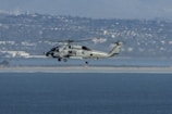 A military helicopter is flying over a body of water with a distant cityscape and mountainous backdrop. The helicopter appears to be hovering at a moderate altitude, and the clear sky suggests good weather conditions.