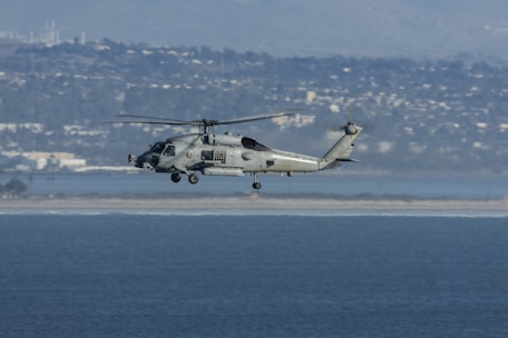 A military helicopter is flying over a body of water with a distant cityscape and mountainous backdrop. The helicopter appears to be hovering at a moderate altitude, and the clear sky suggests good weather conditions.