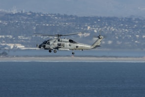 A military helicopter is flying over a body of water with a distant cityscape and mountainous backdrop. The helicopter appears to be hovering at a moderate altitude, and the clear sky suggests good weather conditions.
