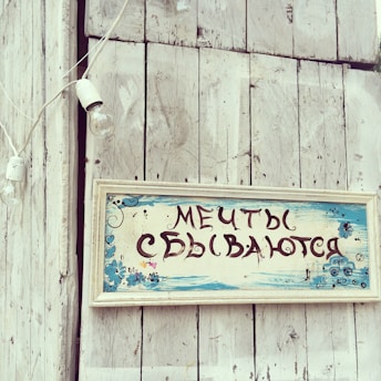 A rustic wooden surface features a vintage-style sign with Russian text, adorned with hand-painted blue flowers and decorative elements. The background includes a whitewashed wooden wall and a hanging string of light bulbs, contributing to a quaint and nostalgic atmosphere.