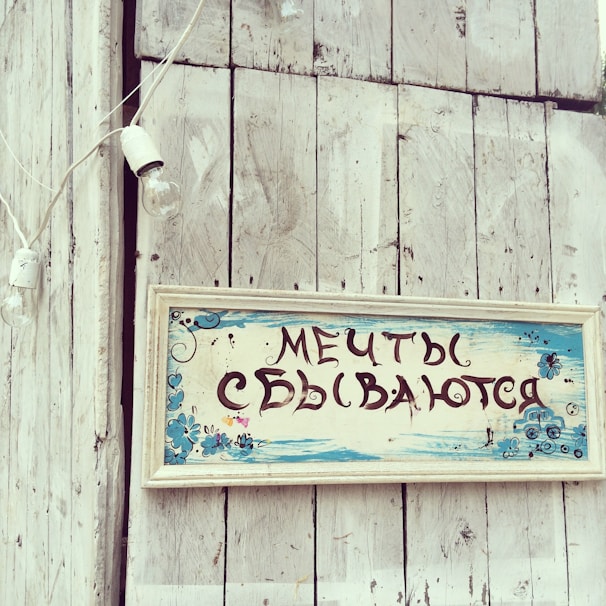 A rustic wooden surface features a vintage-style sign with Russian text, adorned with hand-painted blue flowers and decorative elements. The background includes a whitewashed wooden wall and a hanging string of light bulbs, contributing to a quaint and nostalgic atmosphere.