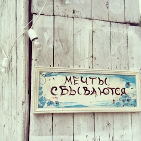 A rustic wooden surface features a vintage-style sign with Russian text, adorned with hand-painted blue flowers and decorative elements. The background includes a whitewashed wooden wall and a hanging string of light bulbs, contributing to a quaint and nostalgic atmosphere.