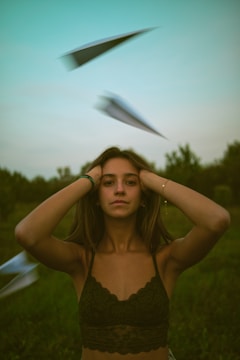 A young woman stands in the foreground with her hands on her head. Paper airplanes fly around her in an outdoor setting with trees visible in the background. The lighting casts a greenish hue over the scene, creating an unusual atmosphere.
